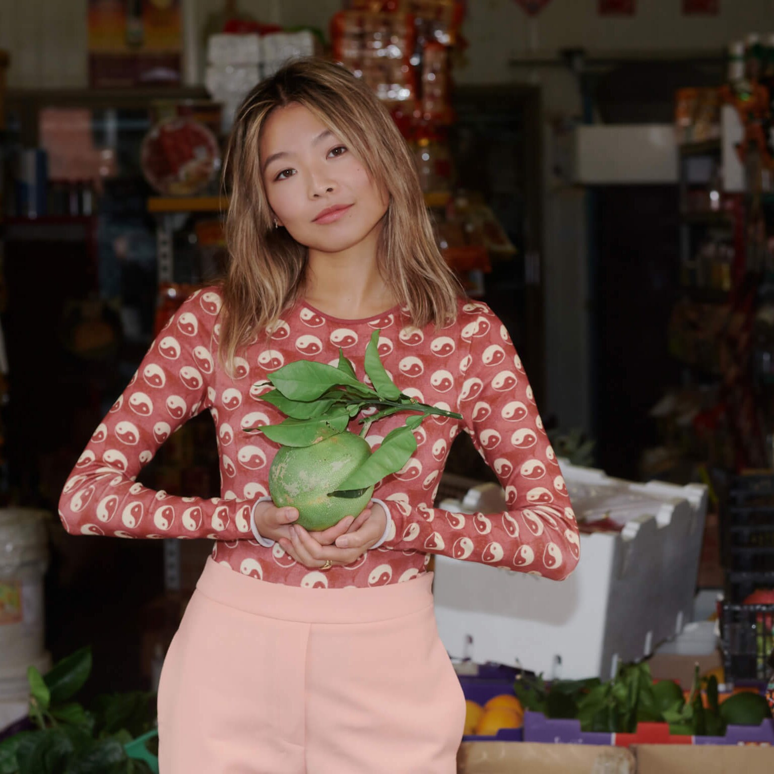 Yan Yan Chan wears a red outfit and holds fruit at an Asian grocery store.