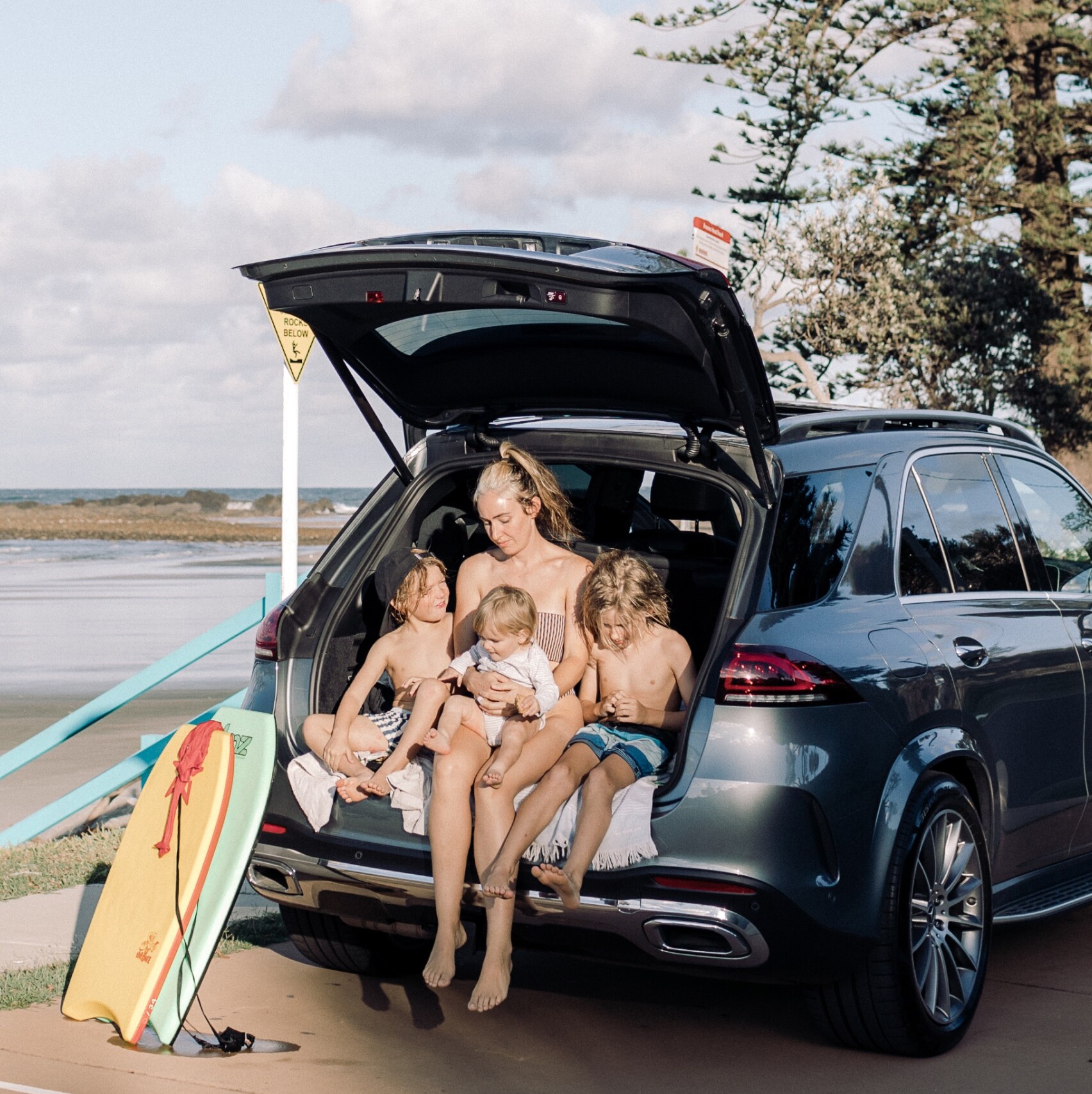 Mother and three children sitting in the boot of a Mercedes-Benz GLE SUV with boogie boards at the beach. Image credit/photographer: Tim Coulson