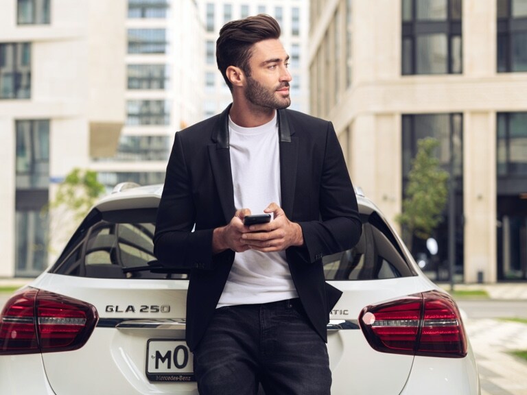 A man leans against his Mercedes-Benz GLA and uses the Mercedes me App on his smartphone and the Navigation Digital Extras to find out what the weather will be like at his destination.