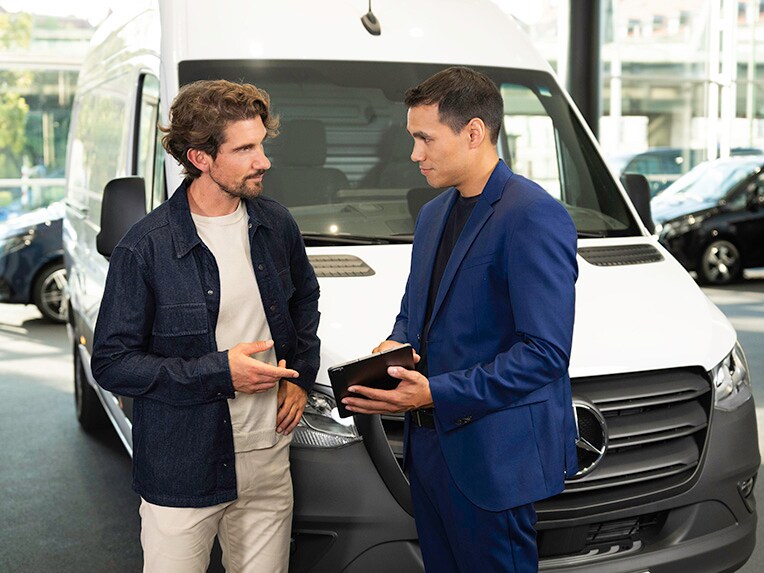 Two people discussing and looking at a mobile device, standing in front of a Mercedes-Benz Van.
