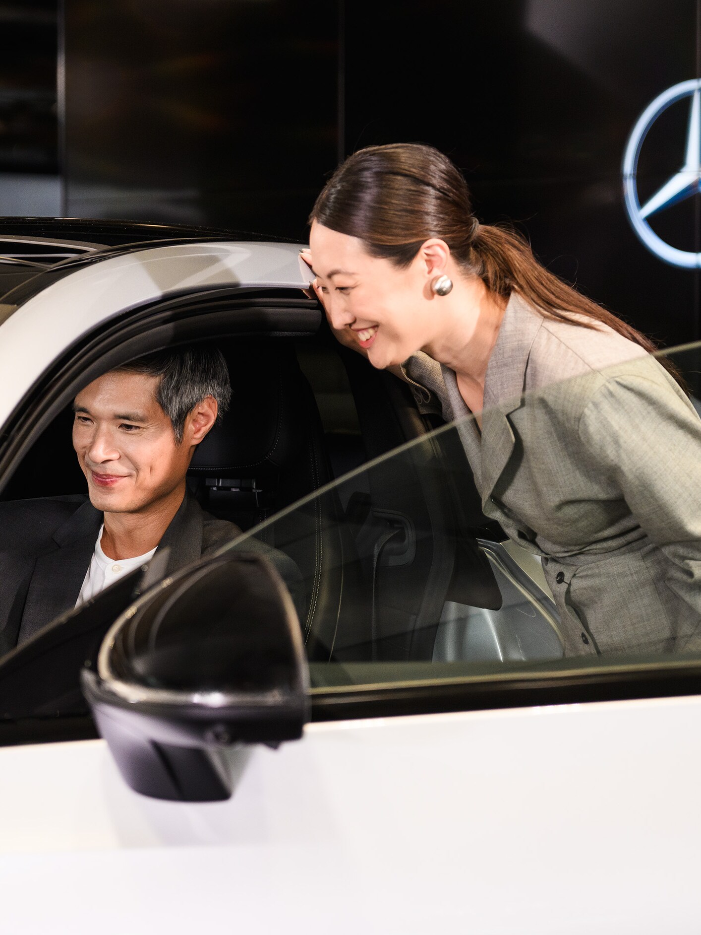 A person sitting in the driver’s seat of a Mercedes-Benz vehicle with someone looking inside the passenger side door open.
