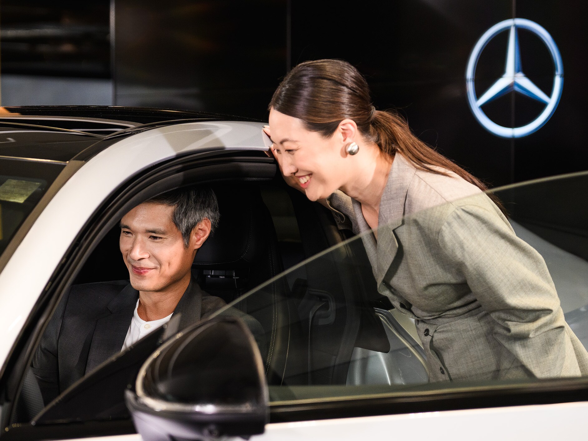A person sitting in the driver’s seat of a Mercedes-Benz vehicle with someone looking inside the passenger side door open.